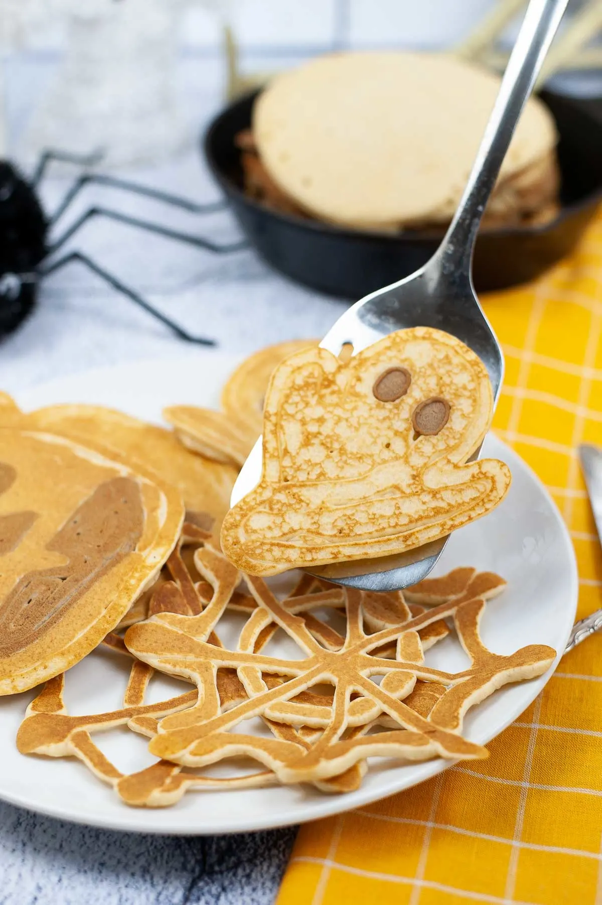 Spatula holding up ghost shaped pancake with other Halloween pancakes on a plate and Halloween decorations.